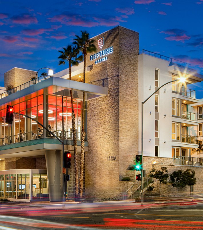 Twilight view of Neptune Marina residential building with illuminated windows and a clear sign, palm trees in the foreground under a dusk sky.