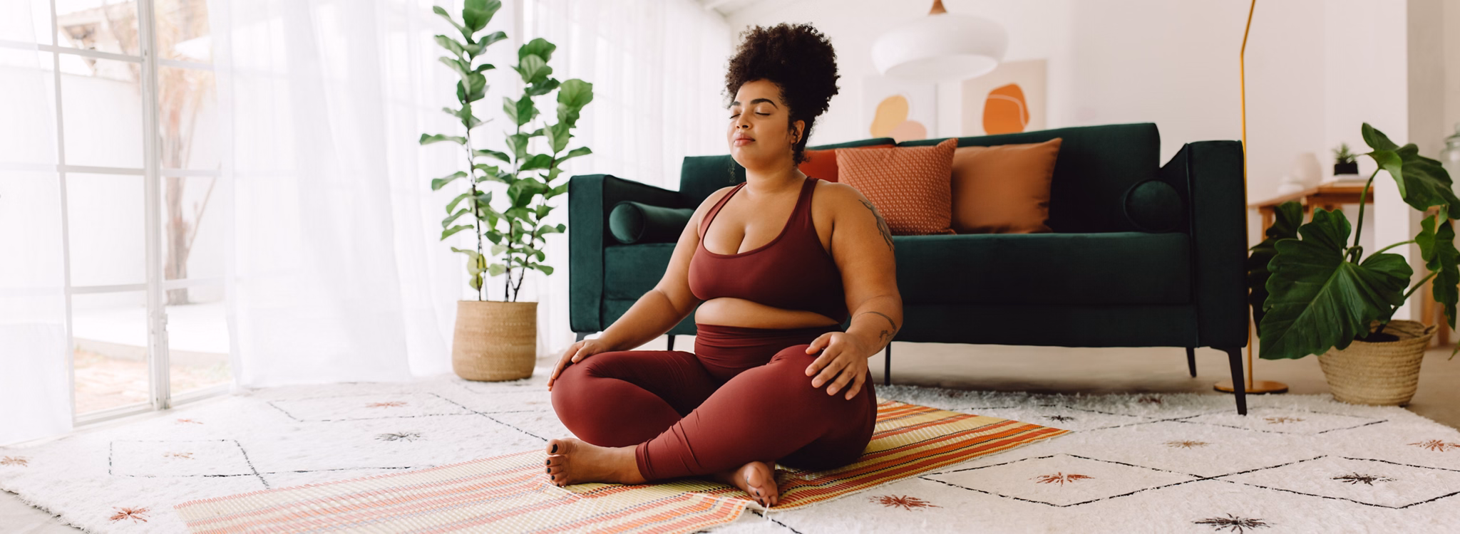 Plus size woman sitting cross legged and closed eyes in living room. Pretty female in fitness wear practicing meditation at home.