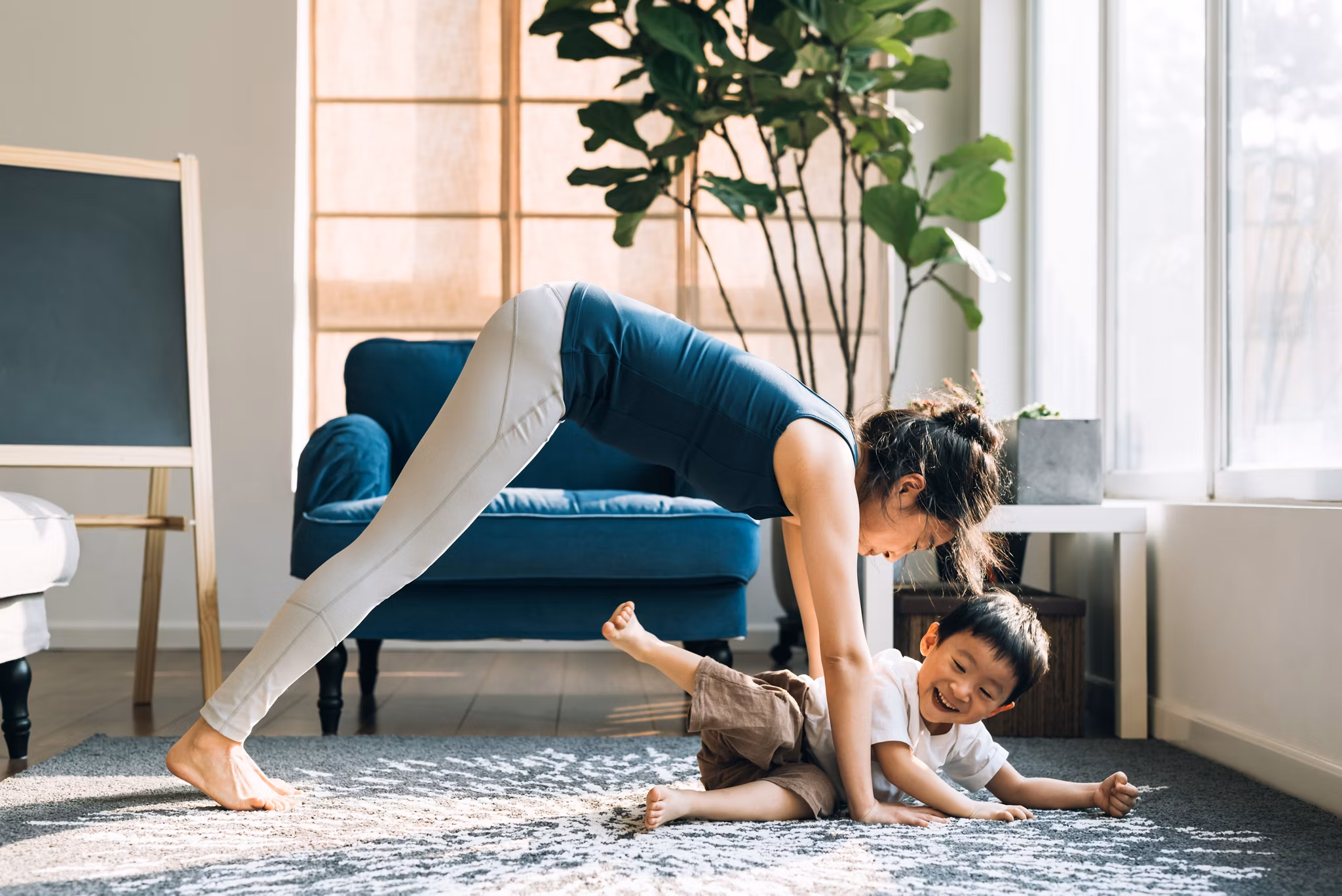 A woman in athletic wear is performing a yoga pose while interacting with a young child in a bright, modern living room