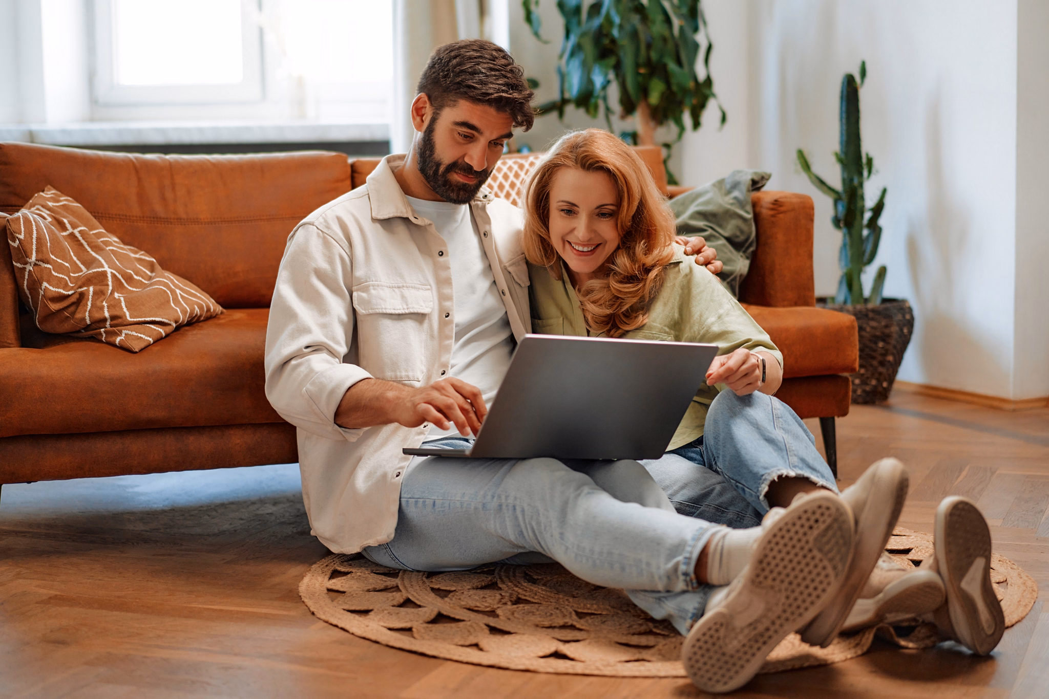 Adult beautiful couple with laptop sitting on the floor near the sofa in the living room, doing online shopping, working or having fun together.