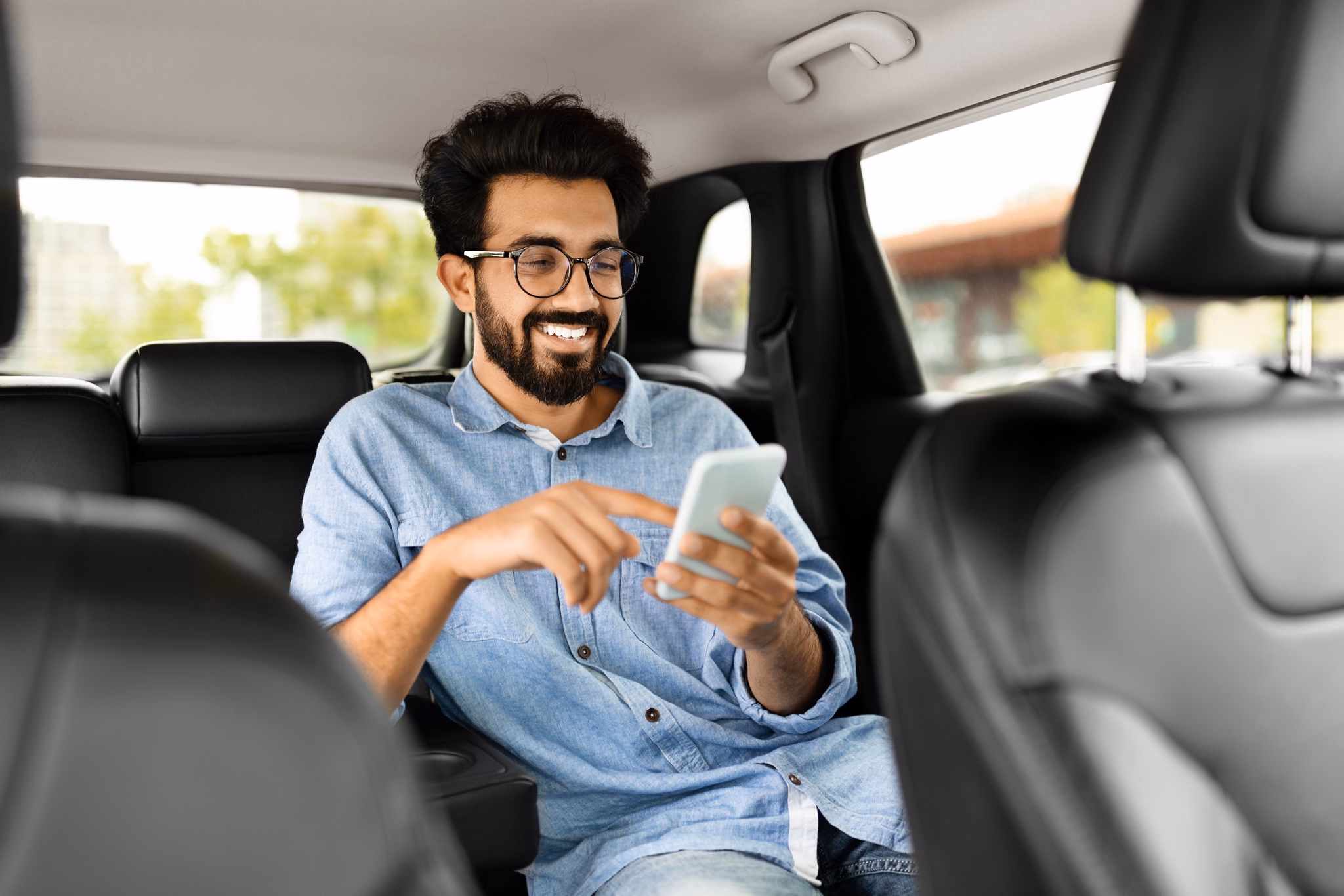 Cheerful handsome millennial indian guy in casual wearing eyeglasses sitting on auto back seat, using smartphone and smiling, hindu man chatting with friends while sitting in taxi