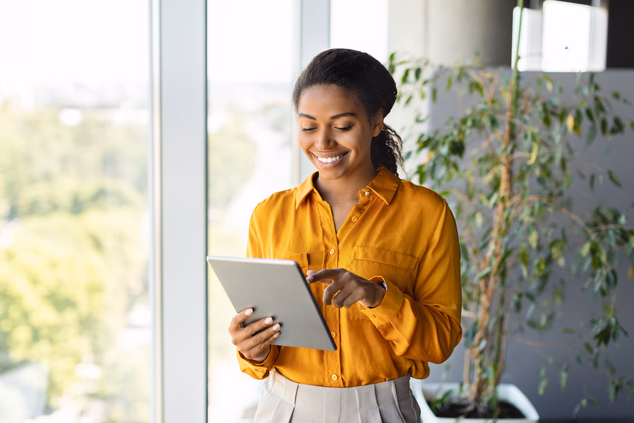 Happy black female entrepreneur using tablet standing near window at modern coworking office, chatting with client. Focused lady browsing internet, free copy space banner mockup