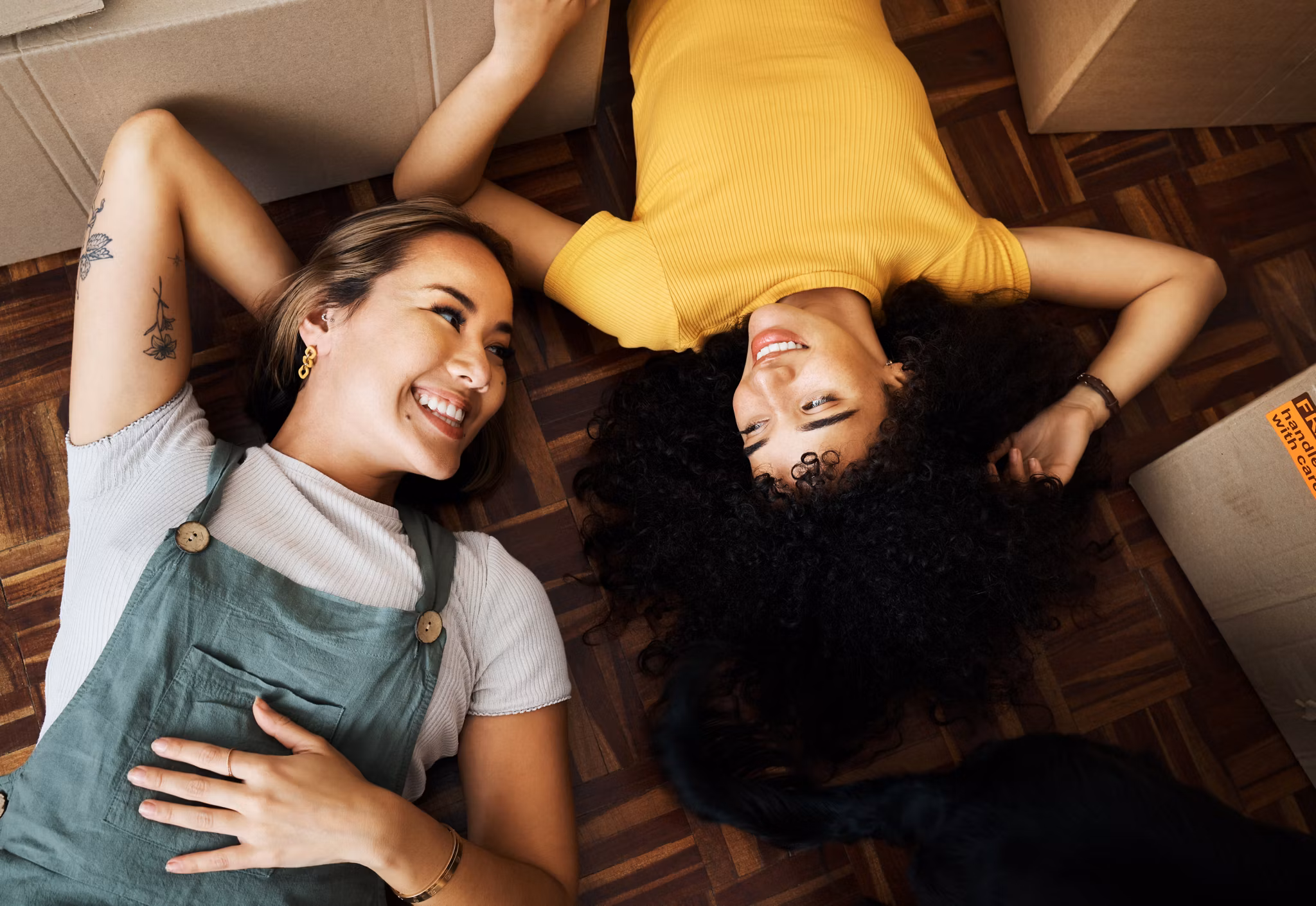 Top view, lesbian and couple with boxes, homeowners and achievement with love, bonding and property. Queer people, lgbtq or happy women on the ground, new apartment and conversation with real estate.