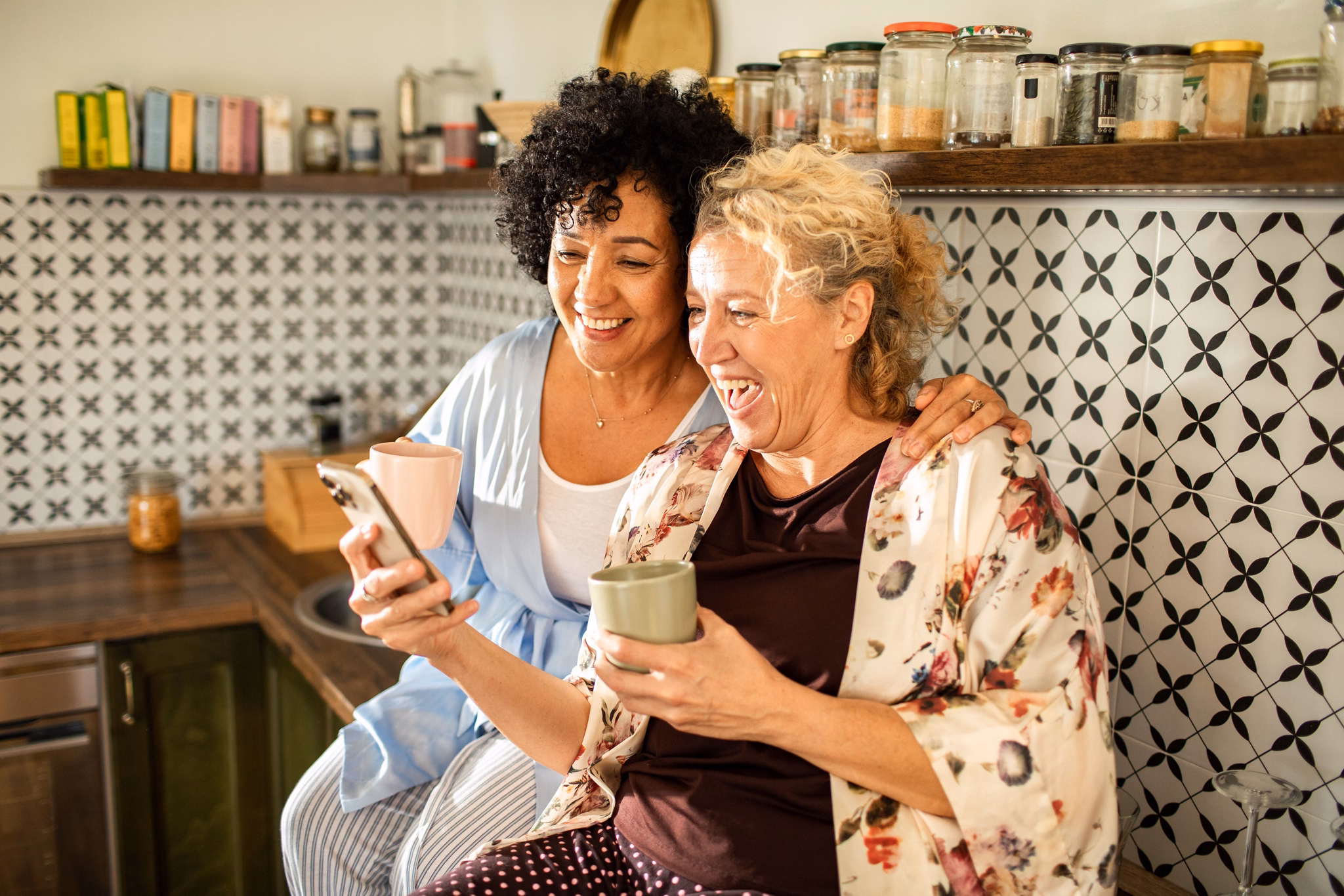 Happy mature lesbian couple laughing together while looking at phone and drinking coffee in kitchen
