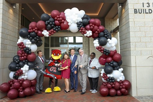 A group of people, including one in a mascot costume, participating in a festive ribbon-cutting ceremony, framed by a decorative balloon arch.
