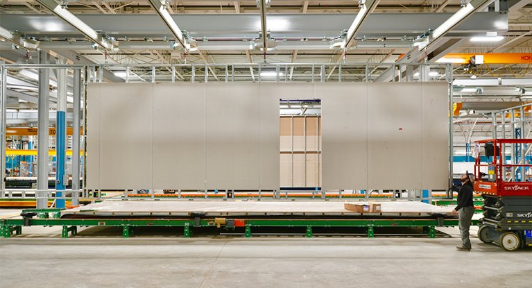 Interior of a manufacturing facility with a worker beside a large panel on an industrial conveyor system under bright lighting.
