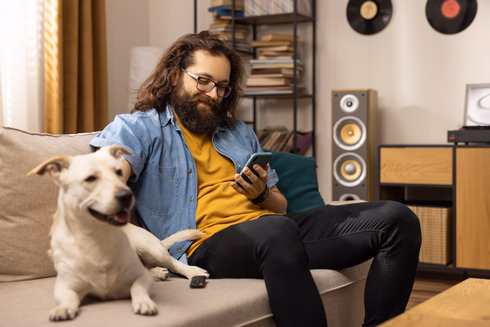 A bearded handsome guy is sitting on the couch with his dog in the living room. The bearded man is browsing insurance policy quotes on his smartphone.