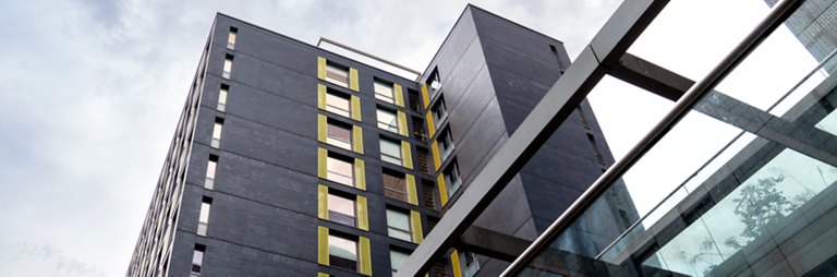 Upward view of a modern building with dark brick facade and yellow window accents under a cloudy sky.