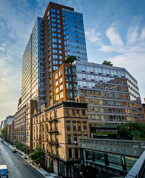 View of a diverse cityscape showing old brownstone buildings in the foreground with a modern, multi-story glass and steel structure in the background against a blue sky.