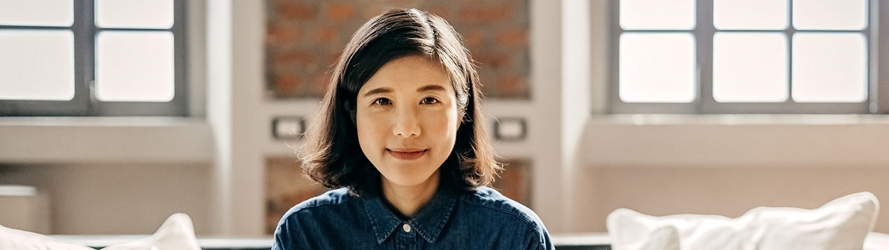 Asian woman with short, black hair sitting on sofa in apartment and looking at camera