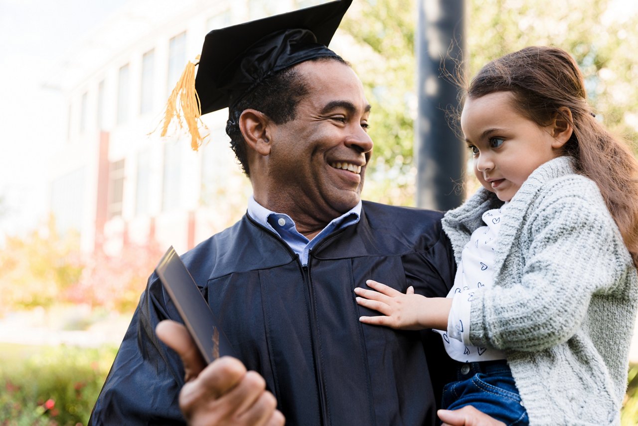 The boomer grandfather, in his graduation gown and mortarboard, carries his cute preschool age granddaughter.  He is happy and proud as he shows her his diploma.