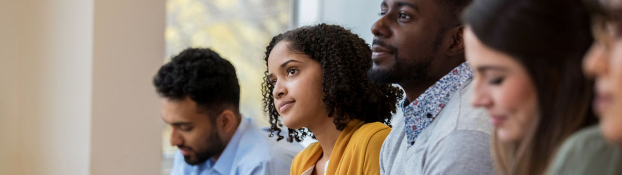 A group of business people sit in a row in a training class. They look at an unseen speaker as they concentrate on his lecture