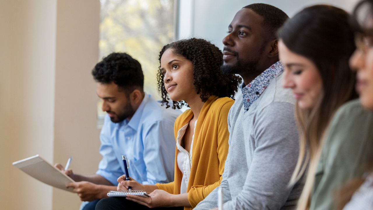 A group of business people sit in a row in a training class. They look at an unseen speaker as they concentrate on the lecture.