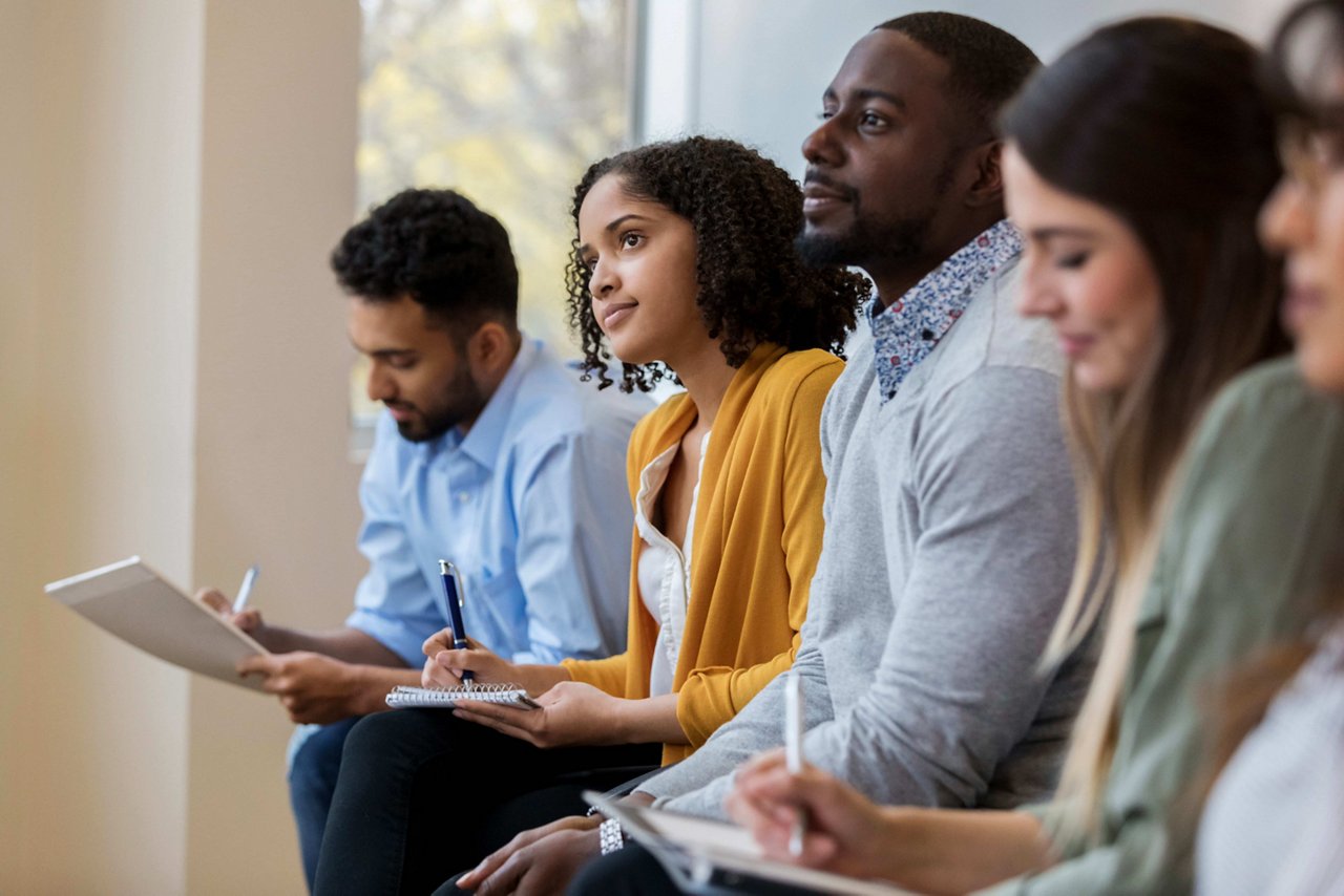 A group of business people sit in a row in a training class. They look at an unseen speaker as they concentrate on the lecture.