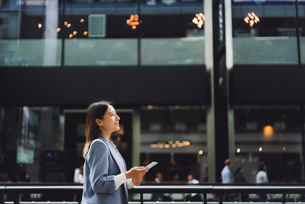 Asian businesswoman walking in a financial area, she is looking up to the sky and holding a smartphone