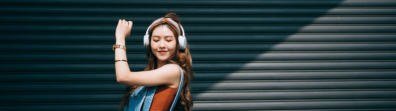 Carefree smiling young Asian woman dancing with her eyes closed while listening to music on headphones outdoors against coloured wall and sunlight. Music and lifestyle