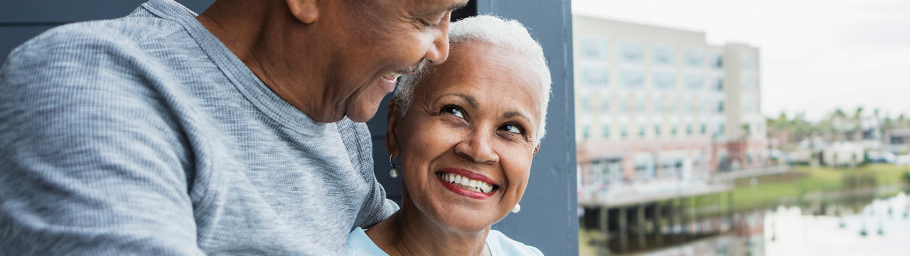 A senior African American couple standing on a balcony overlooking the water. They are looking lovingly into each other's eyes, smiling