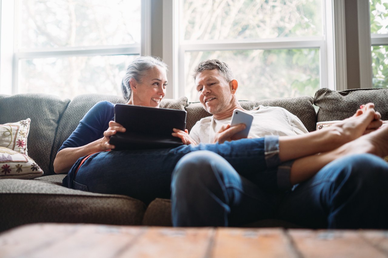 A couple in their 50's relax in their home on the living room couch, enjoying reading and surfing the internet on their mobile touchscreen phones and computer tablet