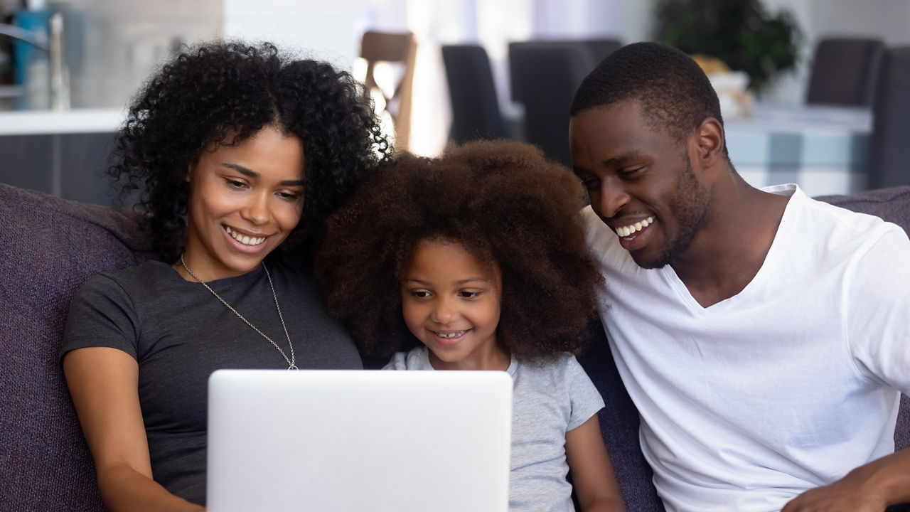Happy African American family with child girl having fun using laptop together at home, black parents and little kid daughter watch funny video, do online shopping, make call look at computer screen