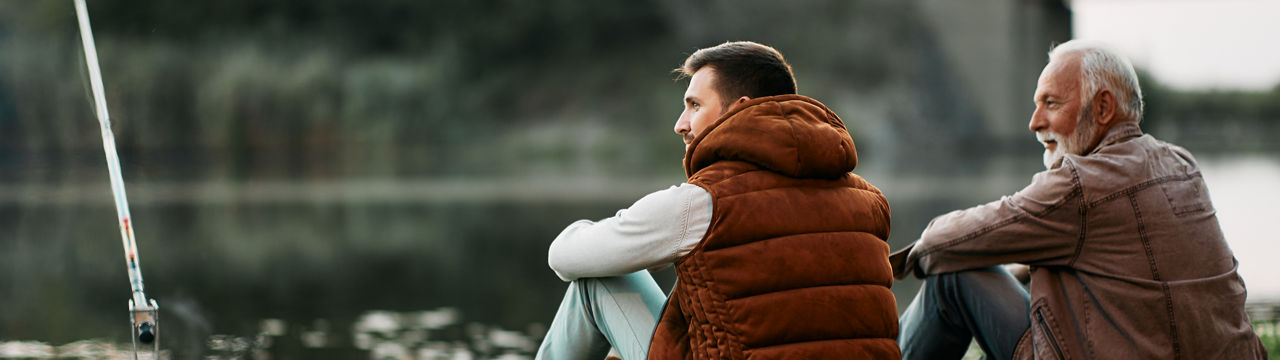 Back view of man and his senior father enjoying while fishing by the river at sunset and looking at the view.