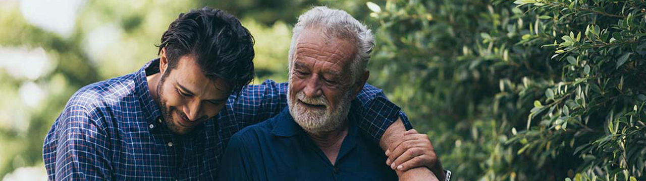 The old man and his son are walking in the park. A man hugs his elderly father. They are happy and smiling.