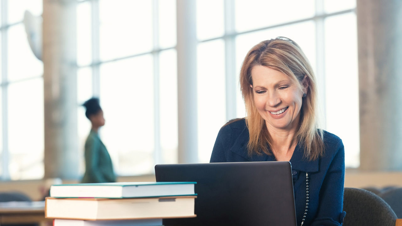 Mature adult Caucasian woman with blonde hair is smiling and looking at laptop computer screen. Adult student or writer is sitting at desk in large modern library with bright windows. She is using a large stack of textbooks to study