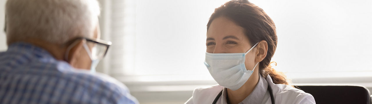 Smiling female doctor in facemask and white uniform talk consult mature male patient at consultation in clinic. Woman GP in facial mask have meeting with elderly client. 