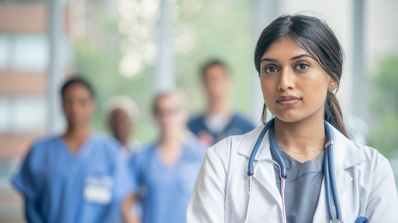 A female doctor stands in the hallway posing for a portrait.  She is wearing a white lab coat and has a stethoscope on as her colleagues stand in the background.