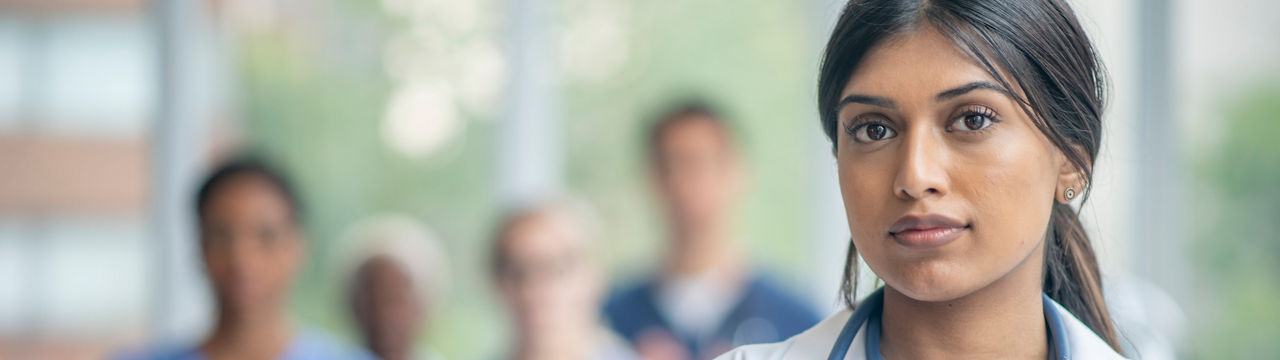 A female doctor stands in the hallway posing for a portrait.  She is wearing a white lab coat and has a stethoscope on as her colleagues stand in the background.