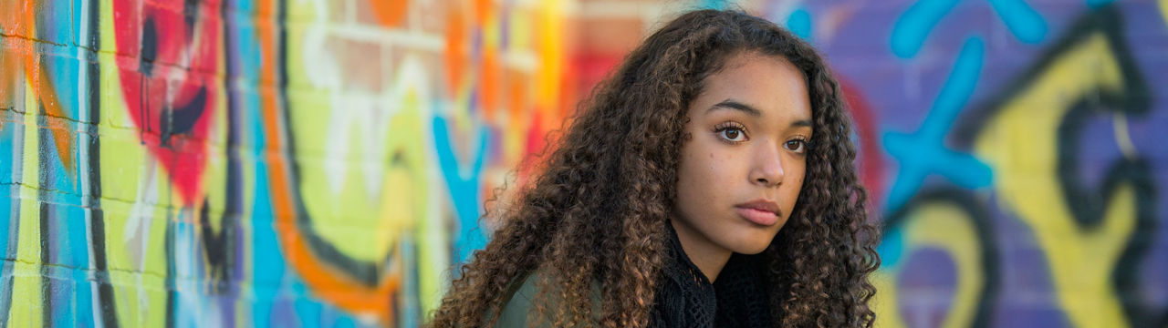 A female teenager of African American descent is sitting alone in front of a graffiti-covered wall. She is looking forward into the distance with a serious expression