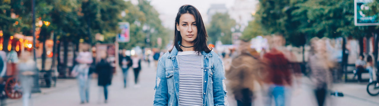 Tired young woman student standing alone in city center and looking at camera with straight face while crowds of men and women are whizzing around.