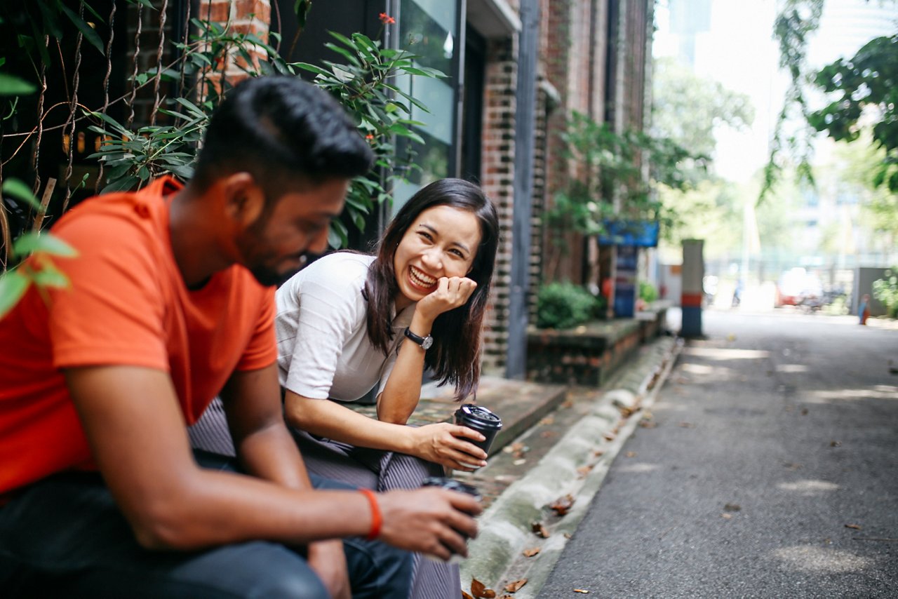 Young people talking and flirting on the streets of Kuala Lumpur, having a coffee break together in front of their office or university library.
