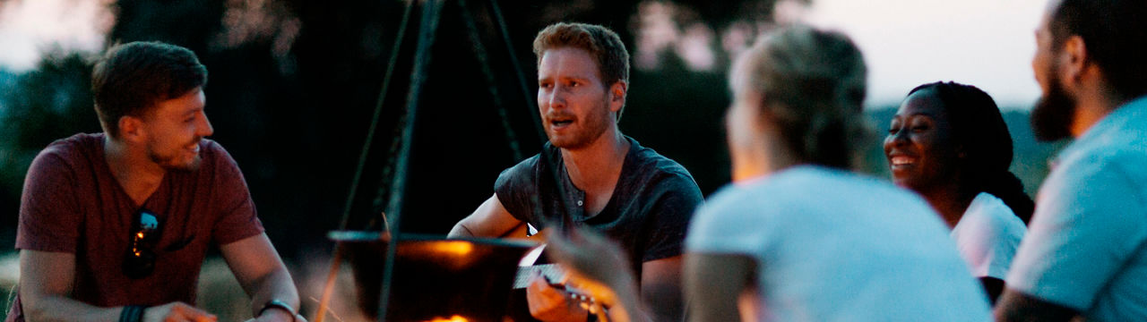 Group of young hikers camping in nature. They enjoy camping and having fun. Beard men playing guitar and singing. They are sitting in front of camping fire