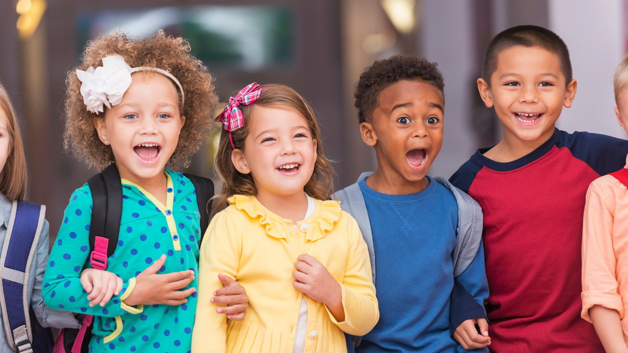 A multi-ethnic group of children standing in a row in a school hallway, excited and laughing, watching something. They are in kindergarten or preschool, carrying bookbags. They are 4 to 6 years old.