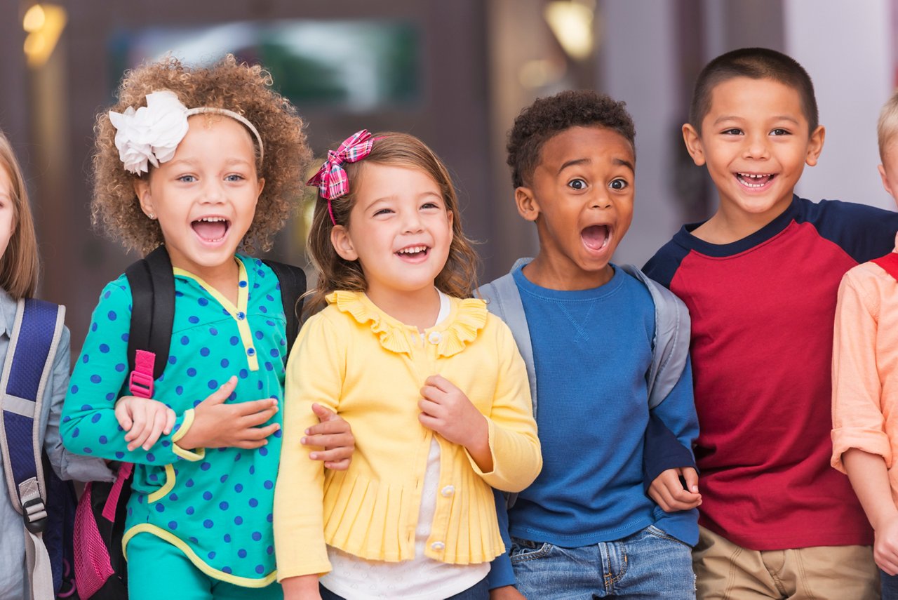 A multi-ethnic group of children standing in a row in a school hallway, excited and laughing, watching something. They are in kindergarten or preschool, carrying bookbags. They are 4 to 6 years old.