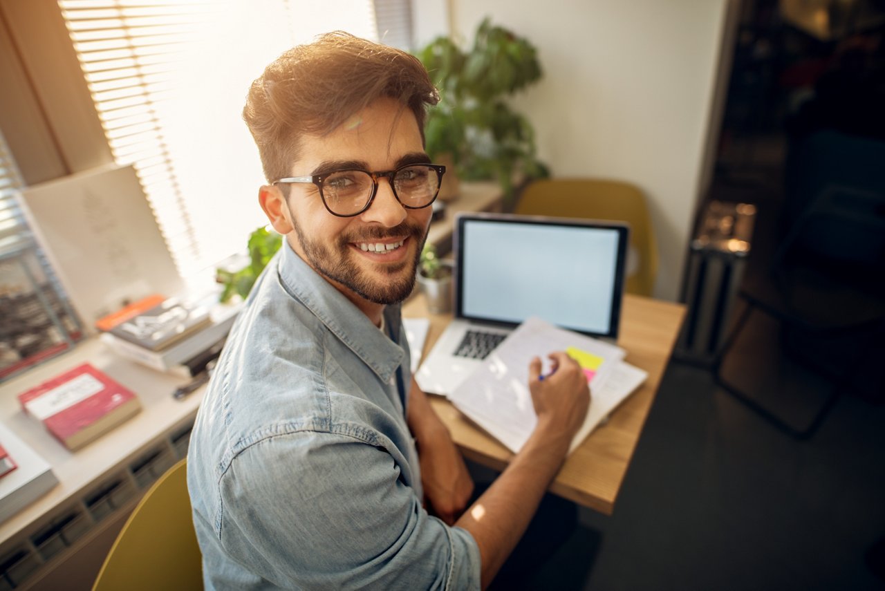 Portrait of happy motivated smiling hipster student learning for a test or an exam at high school library desk while sitting turn backwards and looking at the camera