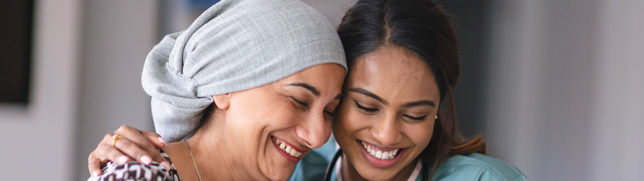 Portrait of an Indian woman fighting cancer with her physician. The doctor is a mixed-race female. The two women are seated next to each other indoors. They are embracing and laughing