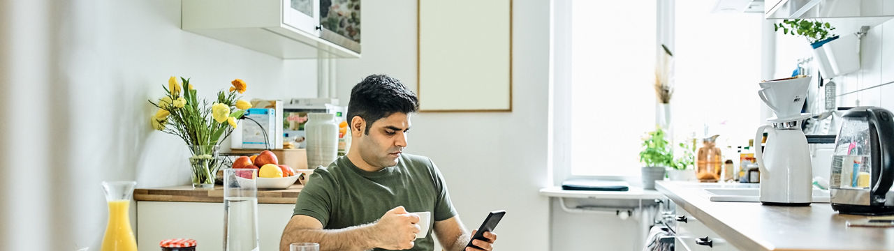Mid adult man drinking coffee and using mobile phone in kitchen. Mid adult male sitting at breakfast tablet with cup of coffee and looking at mobile phone.