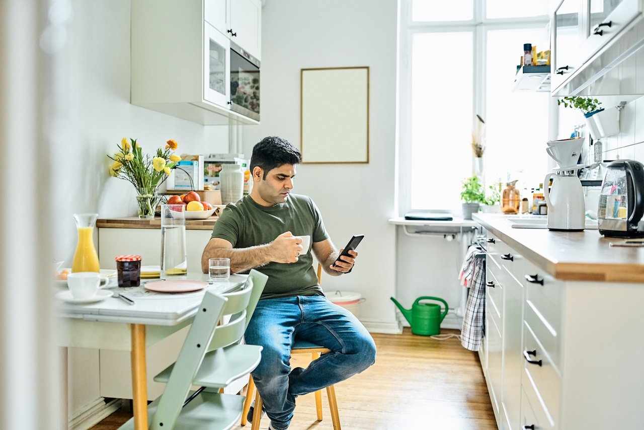 Mid adult man drinking coffee and using mobile phone in kitchen. Mid adult male sitting at breakfast tablet with cup of coffee and looking at mobile phone.