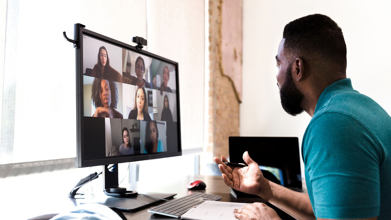 A mid adult man gestures while talking with a group of colleagues during a virtual staff meeting via video conference