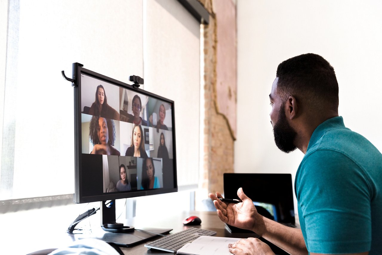 A mid adult man gestures while talking with a group of colleagues during a virtual staff meeting via video conference
