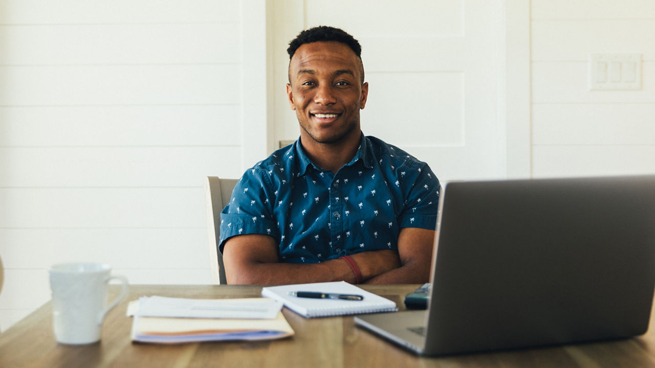 A 30 year old man works on his finances at his kitchen table. He is working on his bank account numbers and preparing to pay his taxes. He is happy, positive and looking forward to better financial times