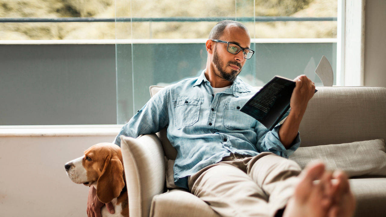 Bearded man comfortably sitting on a coach reading a book and holding his dog .