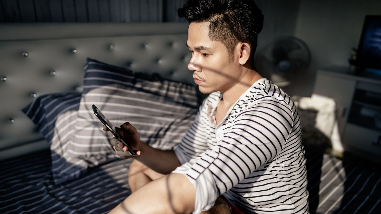 Handsome young Asian man sitting on the bed in the bedroom alone using his smartphone at home