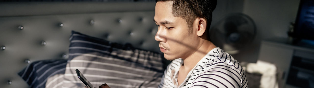 Handsome young Asian man sitting on the bed in the bedroom alone using his smartphone at home