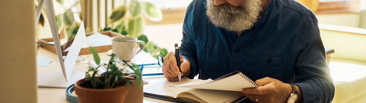 Mature man writing in a notebook diary and sitting alone in a home bedroom. Senior entrepreneur using paperwork and computer to calculate his finances. Journaling poetry or planning business schedule