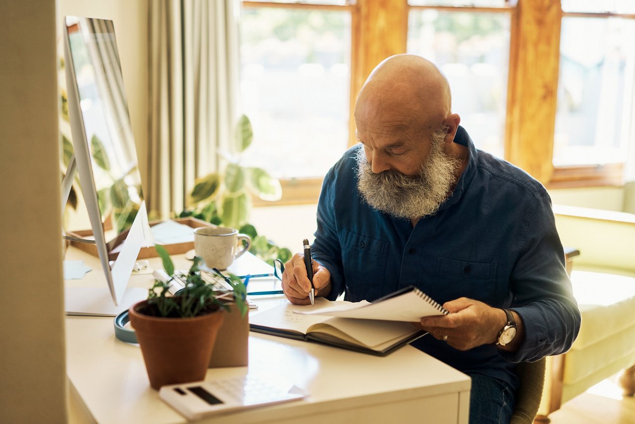 Mature man writing in a notebook diary and sitting alone in a home bedroom. Senior entrepreneur using paperwork and computer to calculate his finances. Journaling poetry or planning business schedule