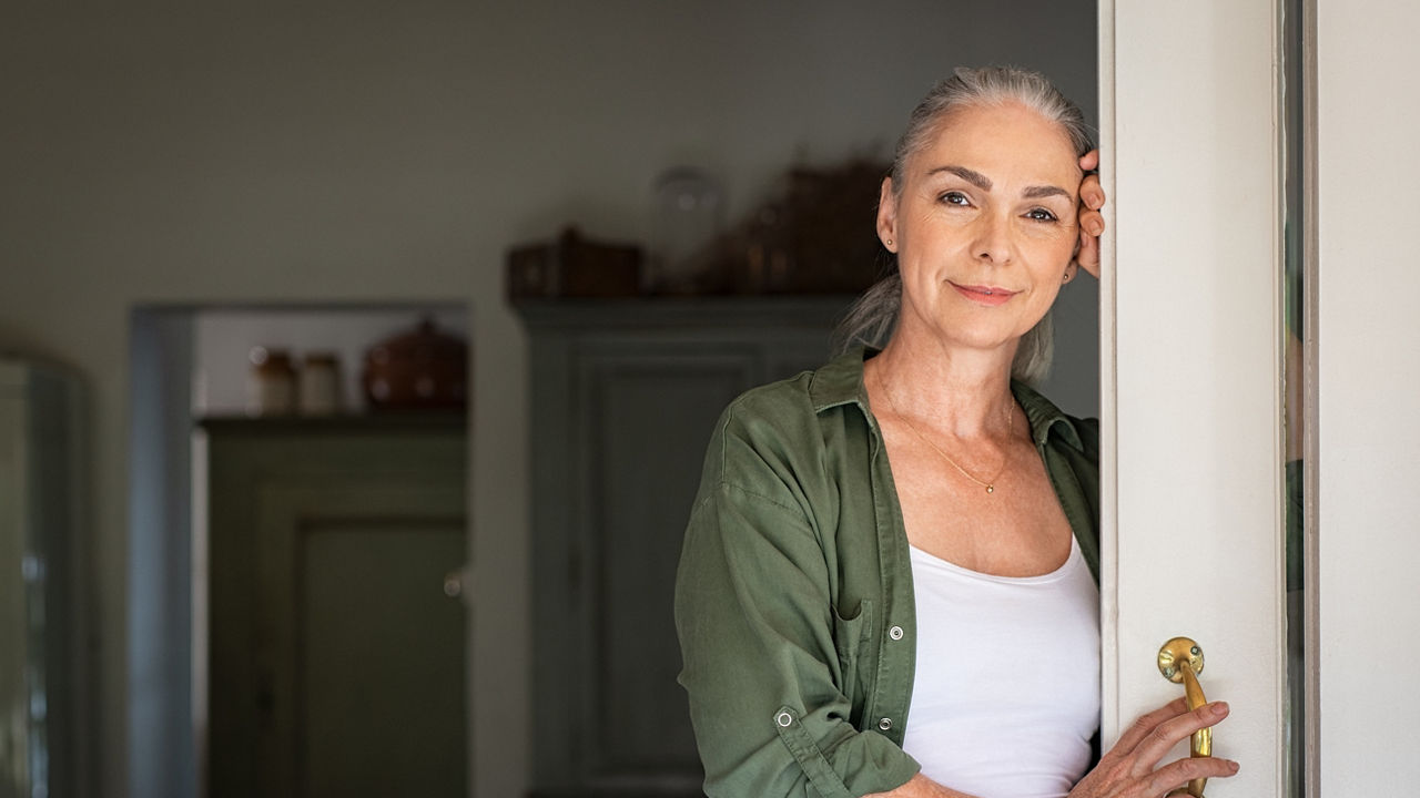Portrait of senior woman standing while leaning against door at home and looking at camera. Smiling mature woman standing at doorway and looking at camera. Portrait of old lady relaxing and thinking with copy space.