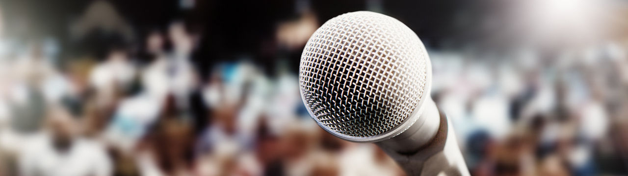 A concert, meeting,  or theatre audience awaits the performer or speaker. An empty mic is in the foreground.