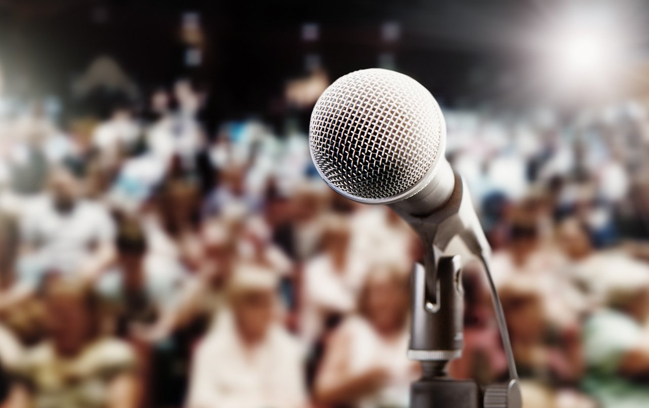 A concert, meeting,  or theatre audience awaits the performer or speaker. An empty mic is in the foreground.